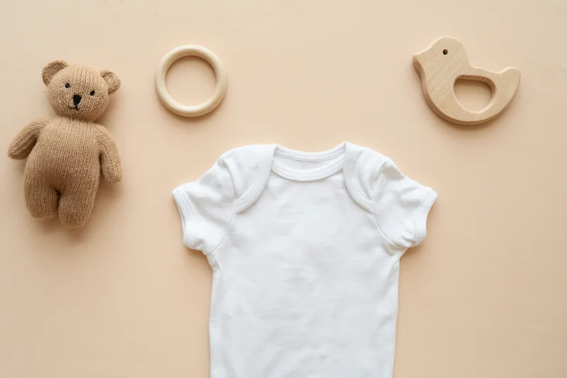 White baby onesie centered on beige background with wooden teether and plush bear toy arranged around it