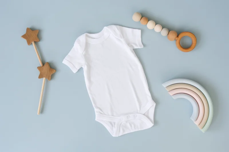 White baby onesie laid flat on soft blue background with wooden toys and rainbow decor