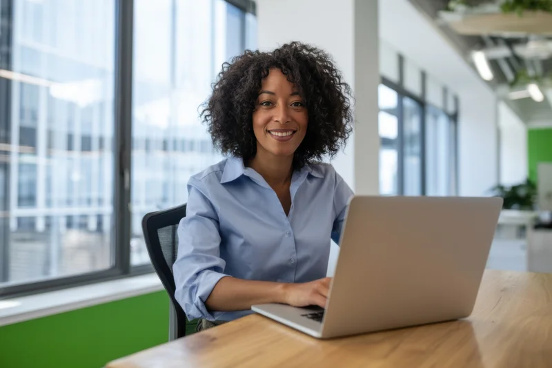 Woman smiling at laptop while working at wooden desk in bright office