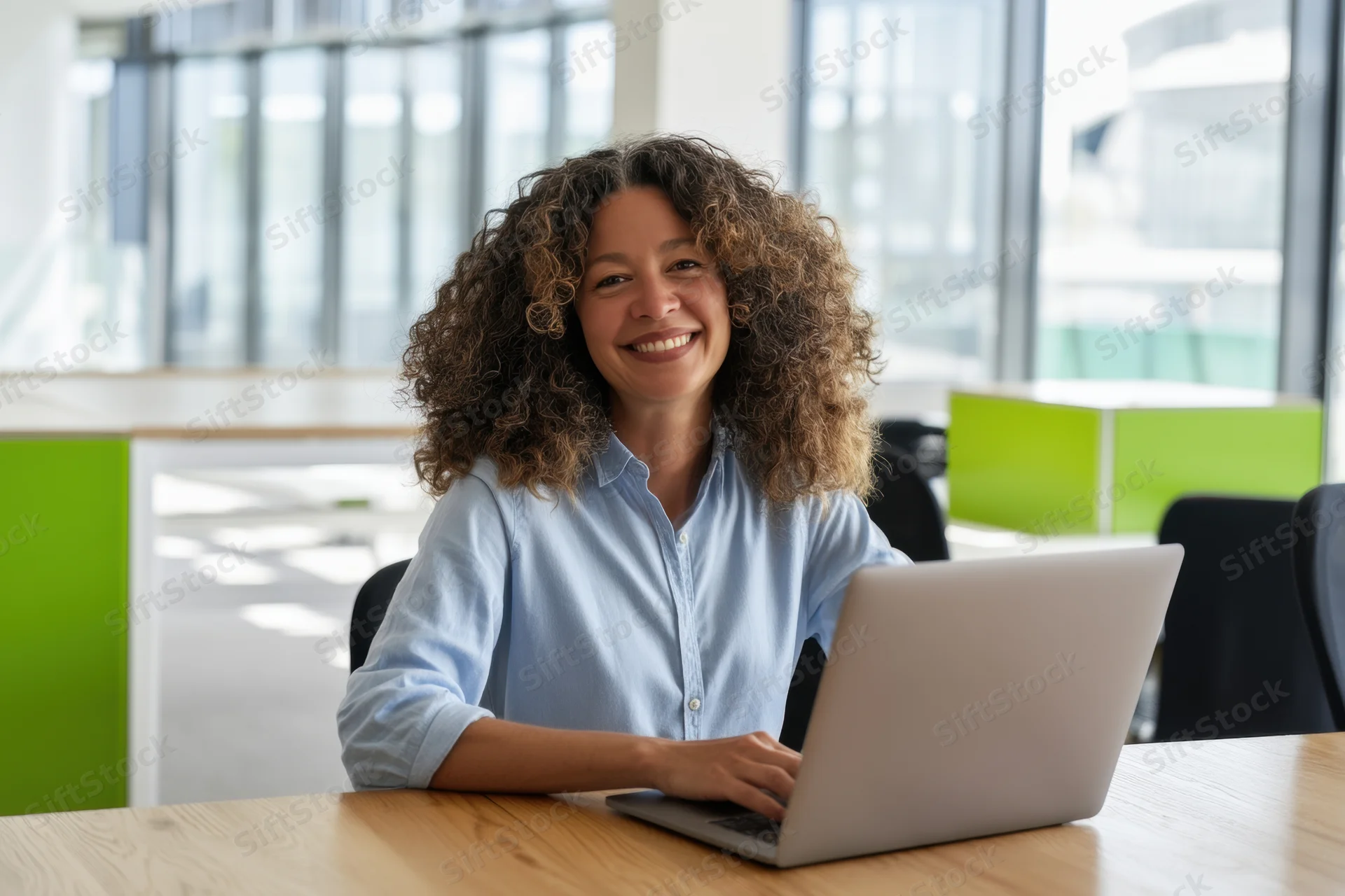 Woman smiling at laptop on wooden desk in bright open office