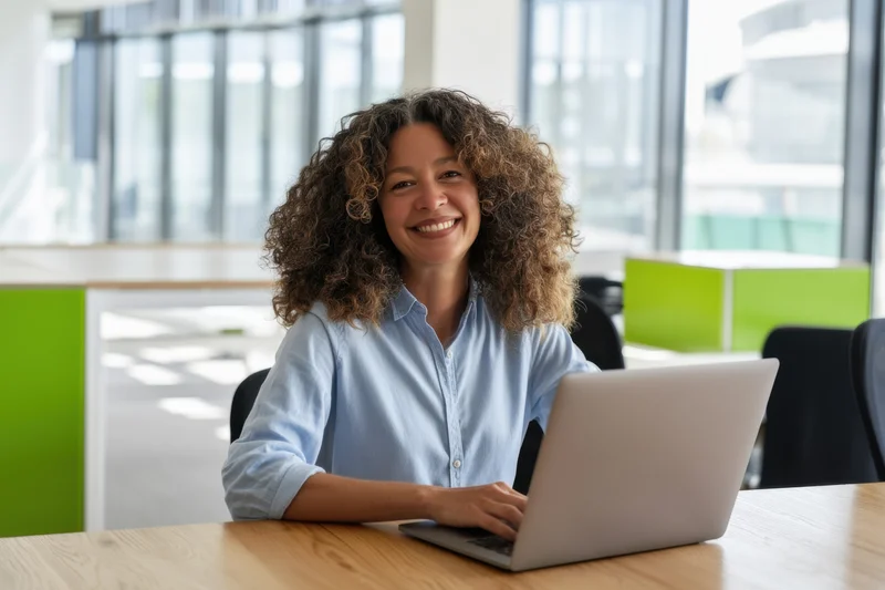 Woman smiling at laptop on wooden desk in bright open office