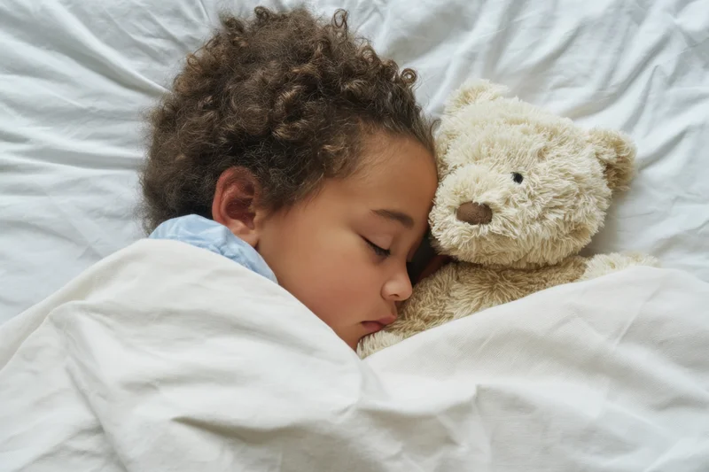 Young child sleeping peacefully in white sheets cuddling a plush teddy bear