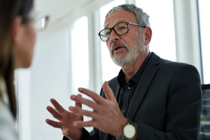 Senior man in dark suit speaking and gesturing during office meeting with woman