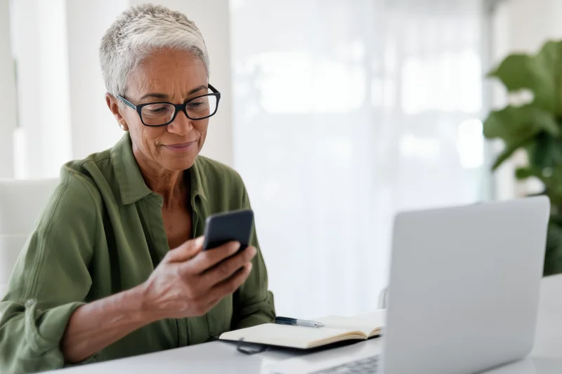 Senior woman with silver hair and glasses smiling while using smartphone at desk with laptop and notebook nearby