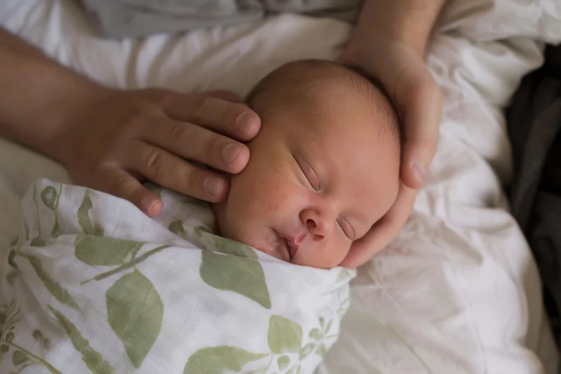 Newborn baby sleeping swaddled in a leaf-patterned blanket with hands gently cradling its head on a bed