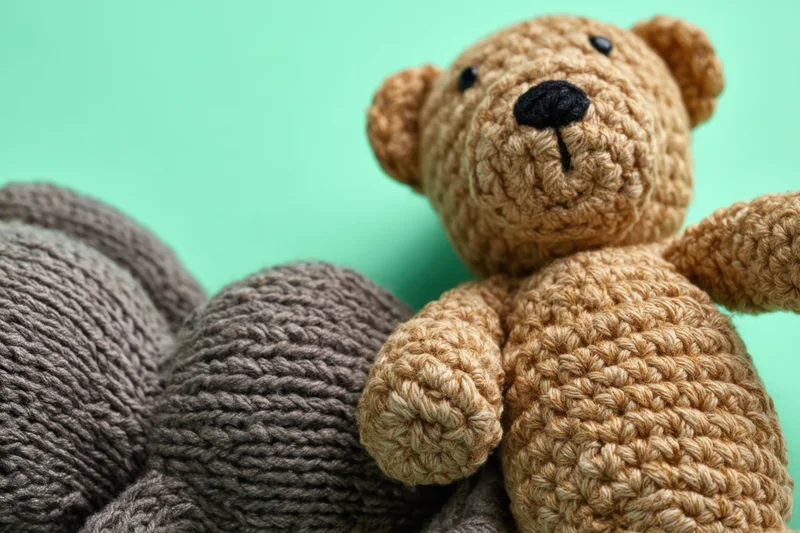 Crocheted teddy bear resting beside gray yarn balls on a green background