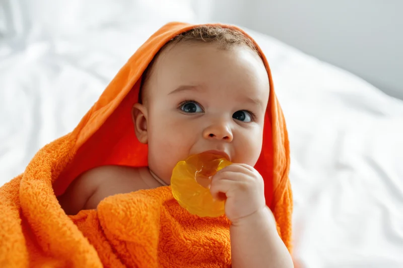 Cheerful infant wrapped in orange hooded towel holding pacifier on white bedding