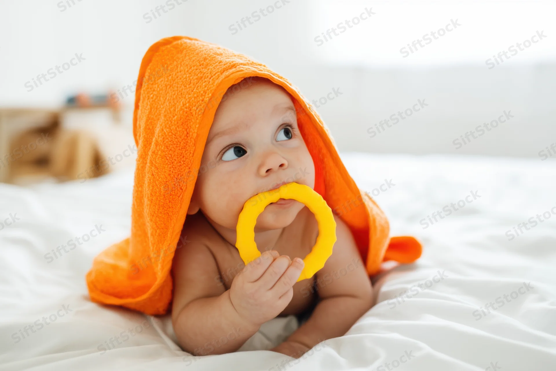 Baby lying on white bed wrapped in orange hooded towel chewing yellow teething ring