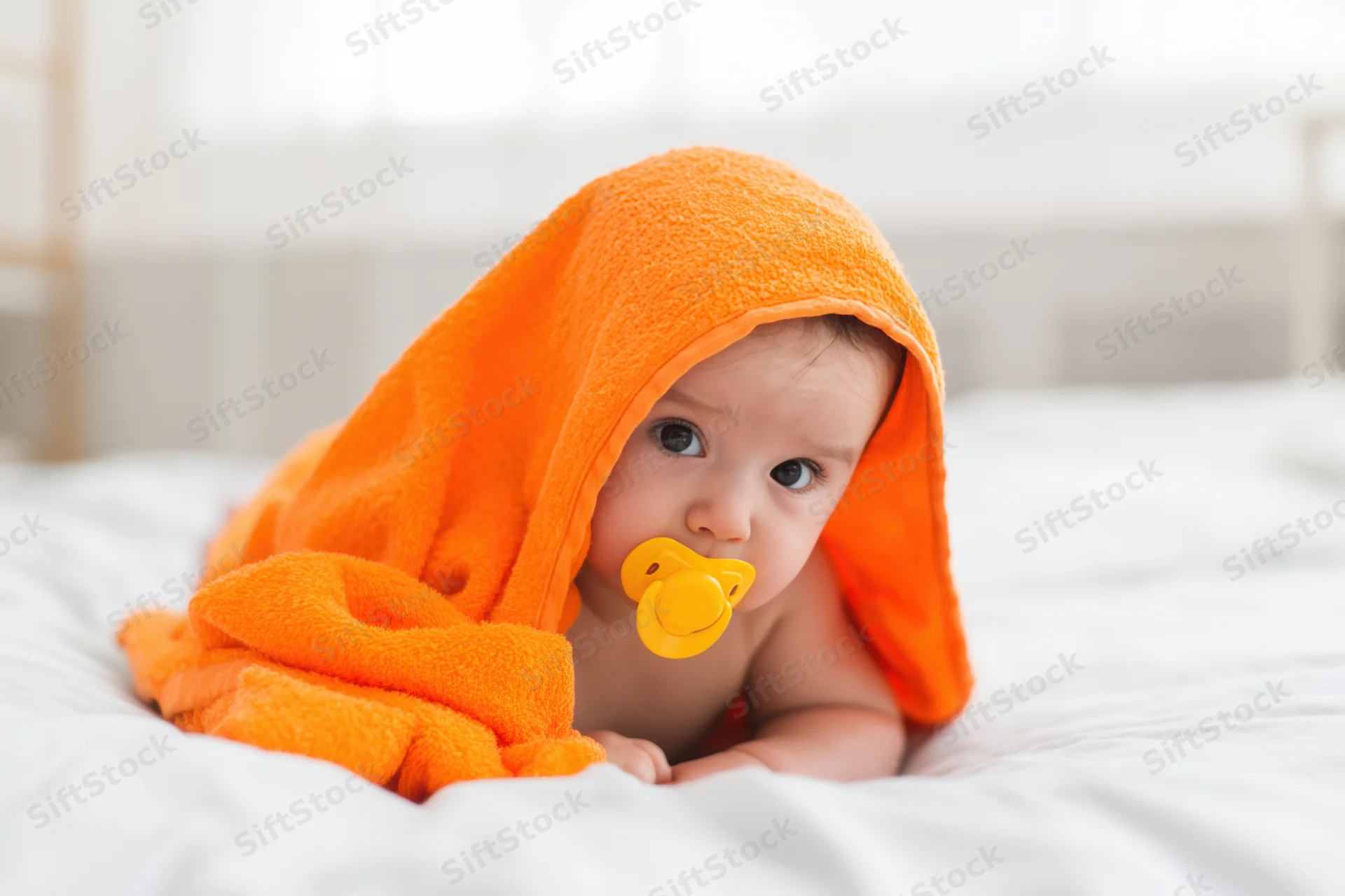 Baby lying on white bedding with orange hooded towel and pacifier in mouth