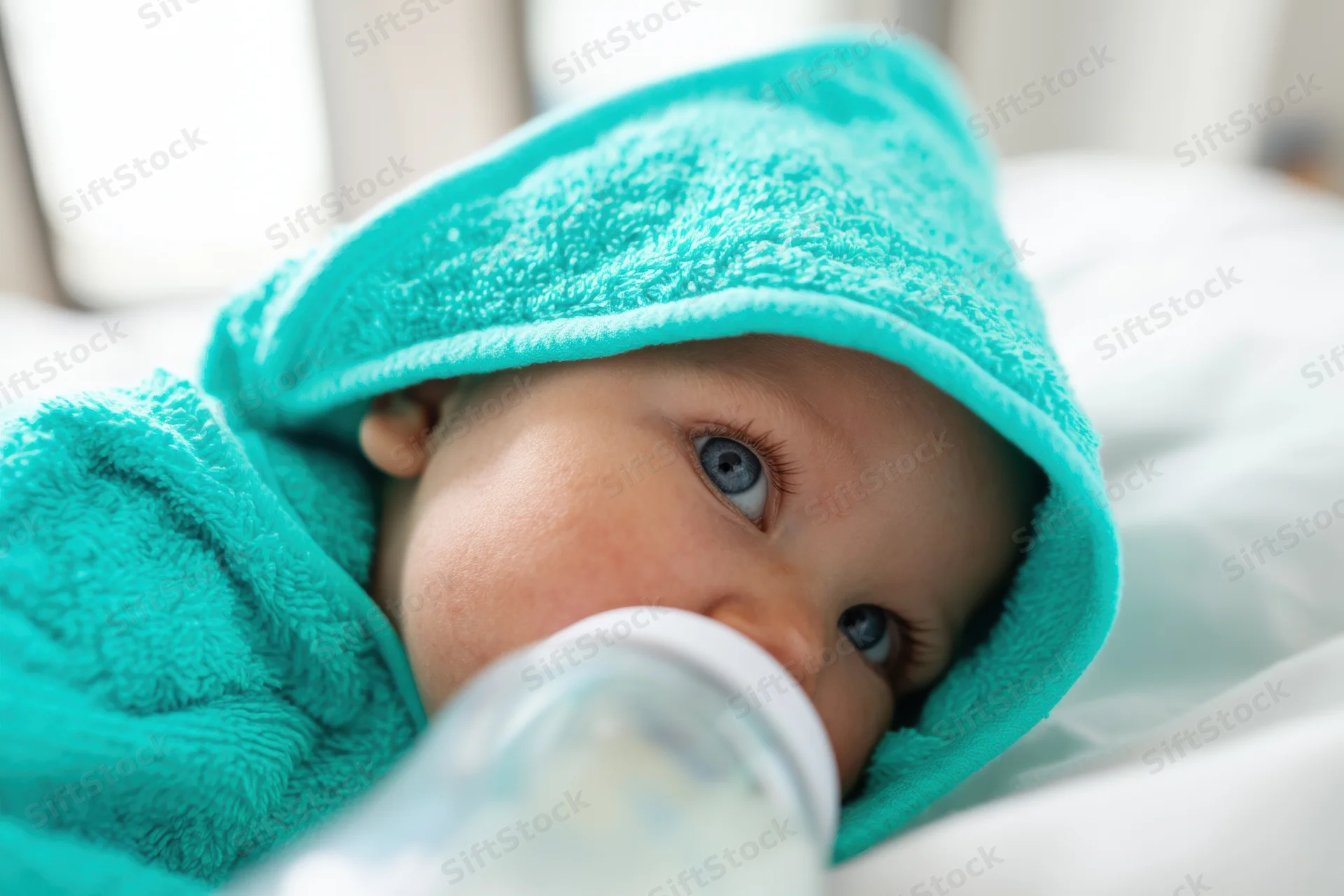 Baby wrapped in turquoise hooded towel lying on white surface with bottle near mouth