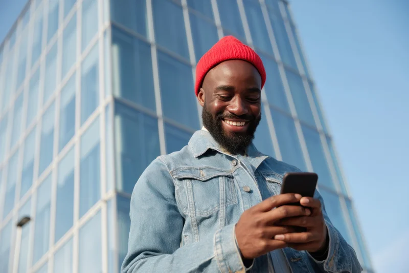 Bearded man in red beanie and denim jacket smiling while using smartphone outdoors near glass skyscraper