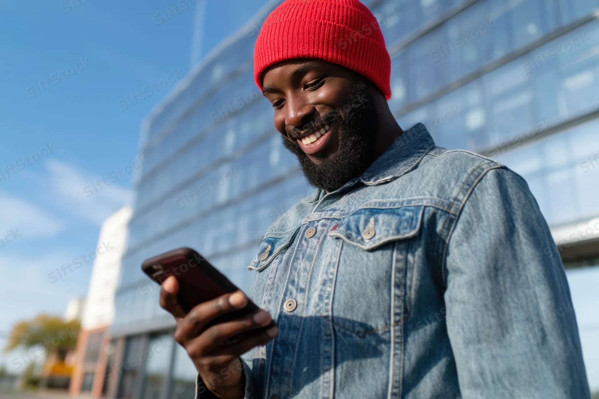 Bearded man in red beanie and denim jacket smiling while using smartphone outdoors near modern glass building