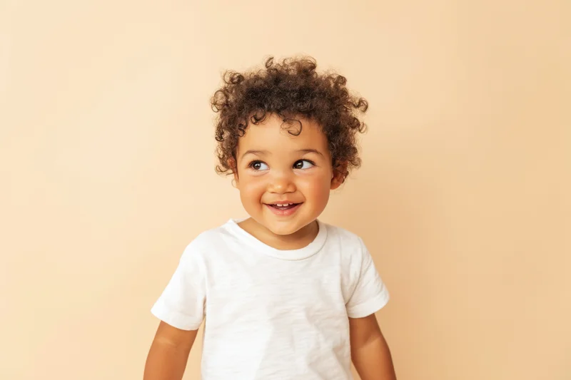 Cheerful toddler with curly hair smiling while wearing a plain white t-shirt against a beige background