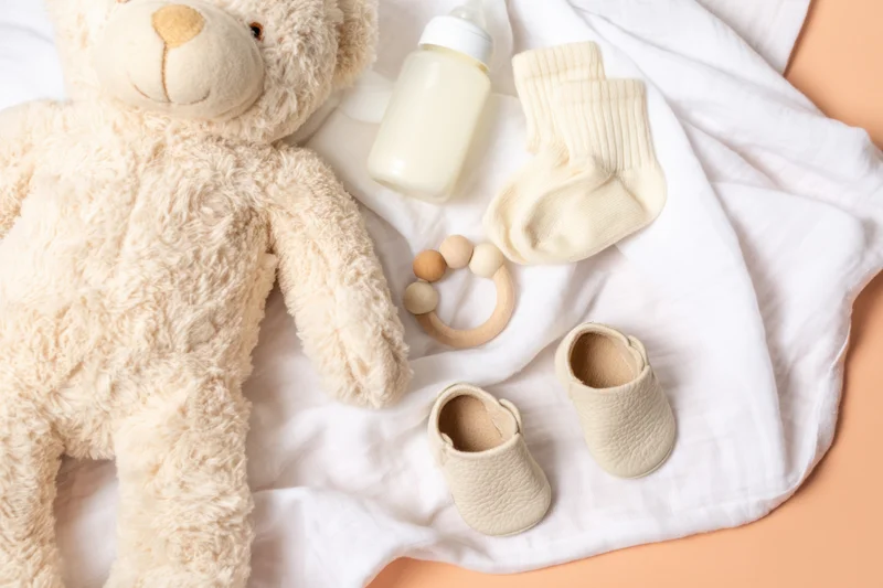 Baby essentials arranged in flat lay on soft white blanket with plush teddy bear and feeding bottle