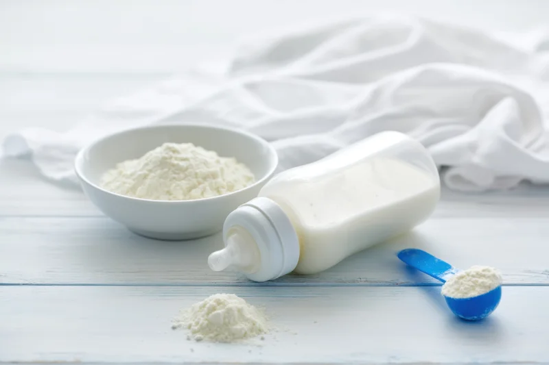 Baby bottle filled with milk lying on pale wooden surface beside bowl of formula powder and blue spoon