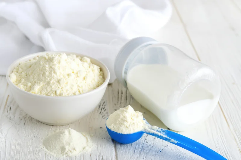 Baby bottle filled with milk lying on a pale wooden surface beside a bowl of infant formula powder and a blue scoop