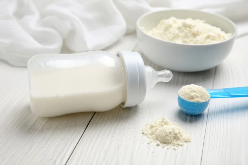 Baby bottle filled with milk lying on a pale wooden surface beside a bowl of infant formula and a scoop of powder