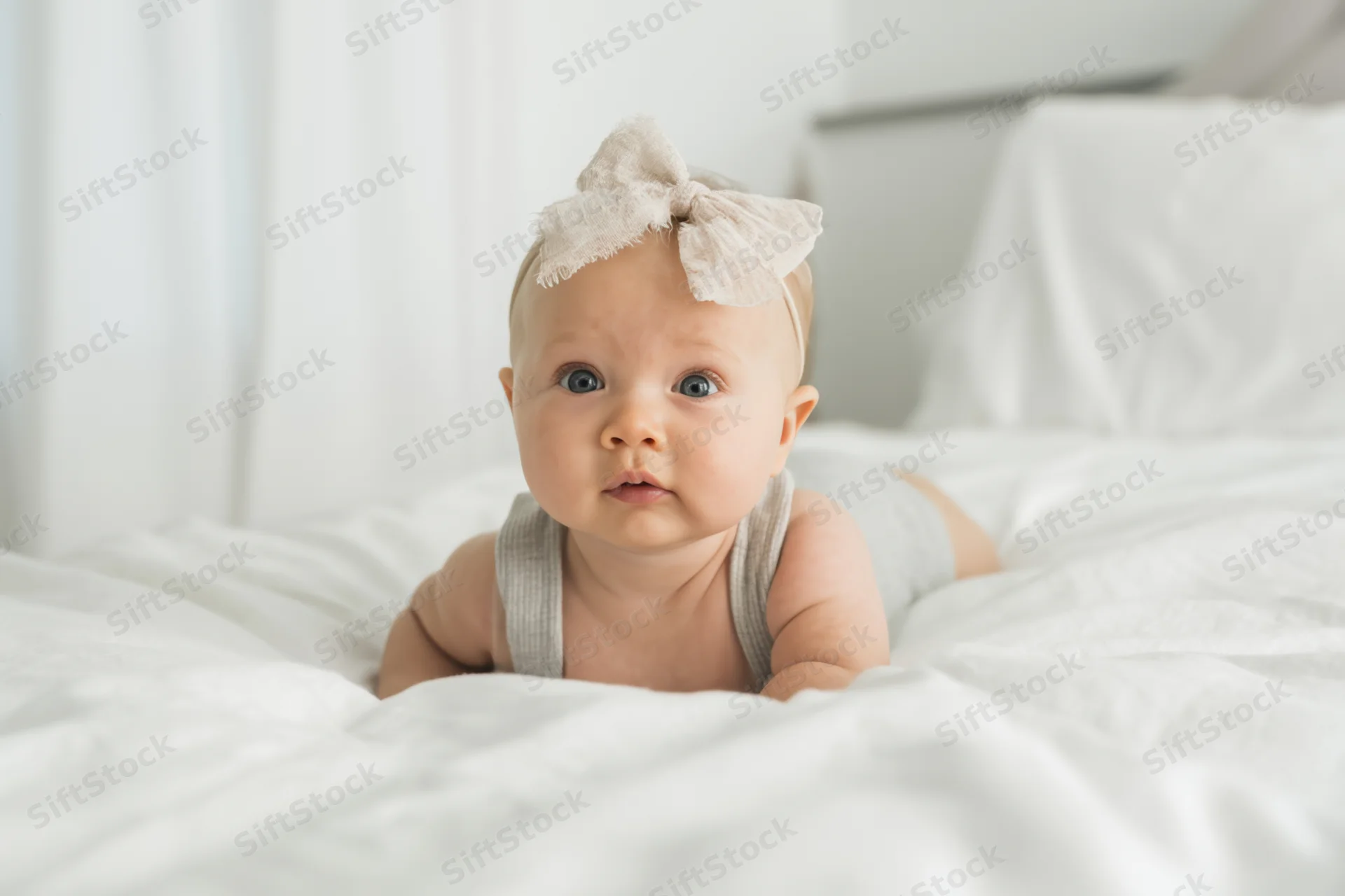 Baby girl lying on stomach on white bed wearing headband and gray tank top