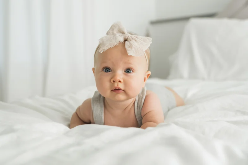 Baby girl lying on stomach on white bed wearing headband and gray tank top