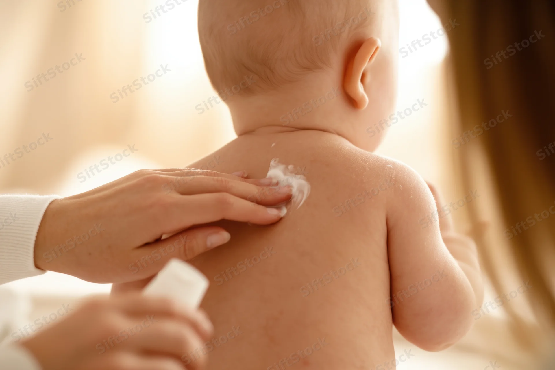 Baby receiving lotion application on back by caregiver at home