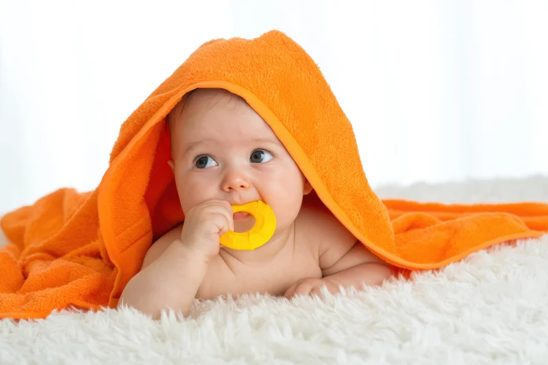 Baby lying on white fluffy blanket wrapped in orange towel holding yellow teether