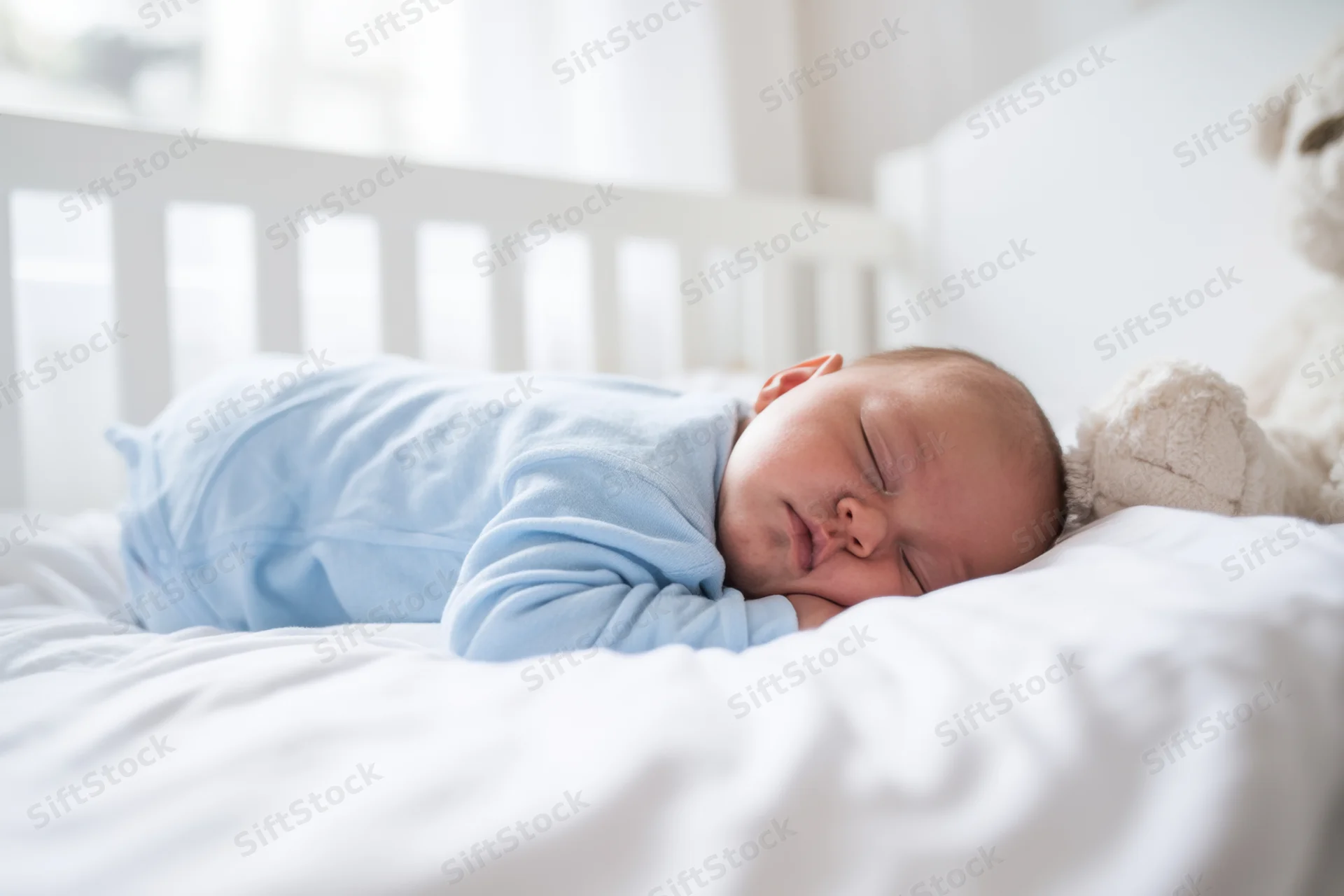 Baby sleeping on white bedding in a crib with a plush teddybear nearby