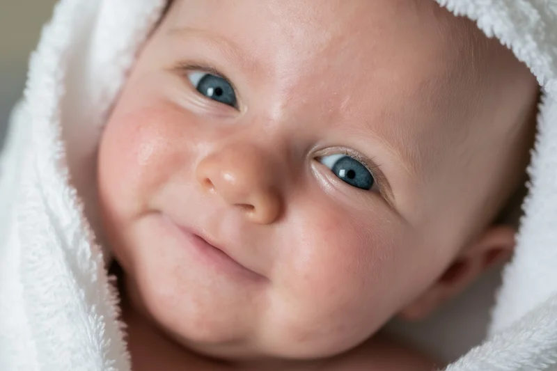 Baby with bright blue eyes peeking from a fluffy white towel, smiling softly in a close-up portrait
