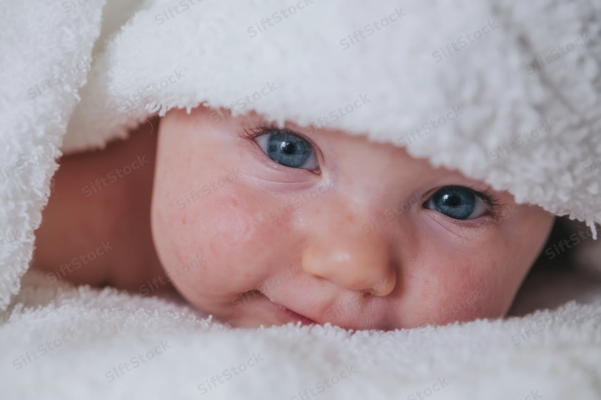 Baby peeking from under a fluffy white towel with bright blue eyes and a slight smile