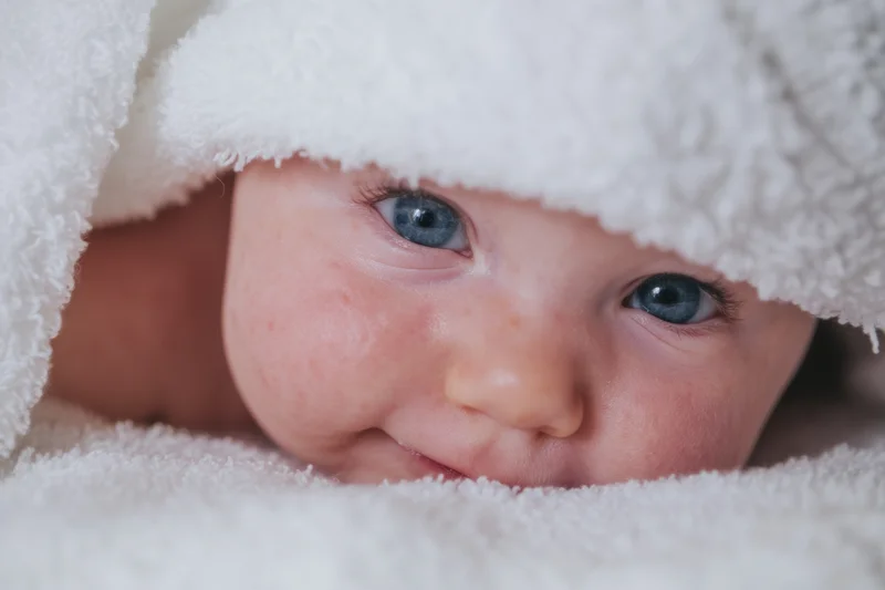 Baby peeking from under a fluffy white towel with bright blue eyes and a slight smile