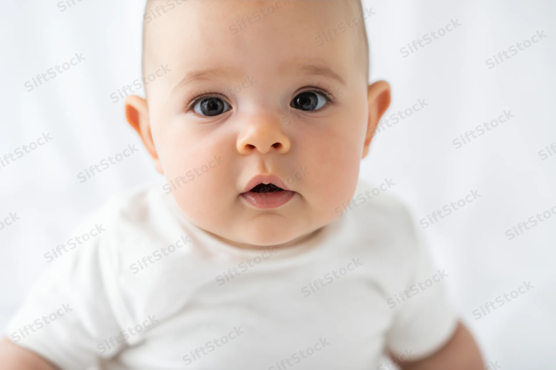 Baby with large dark eyes looking directly at camera in white shirt against plain background
