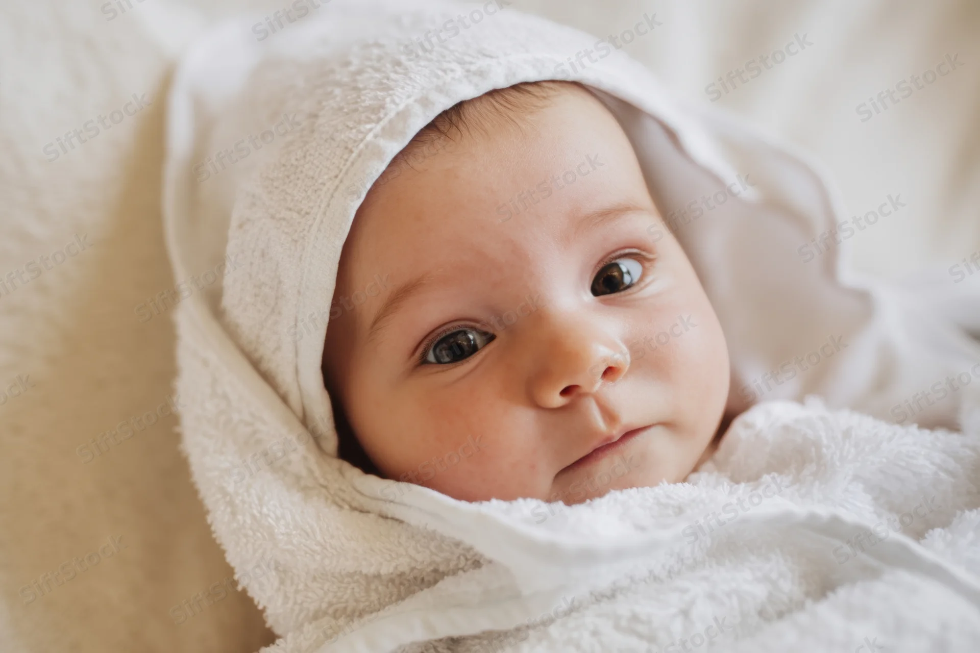 Newborn baby wrapped in a fluffy white hooded towel gazing softly at the camera indoors