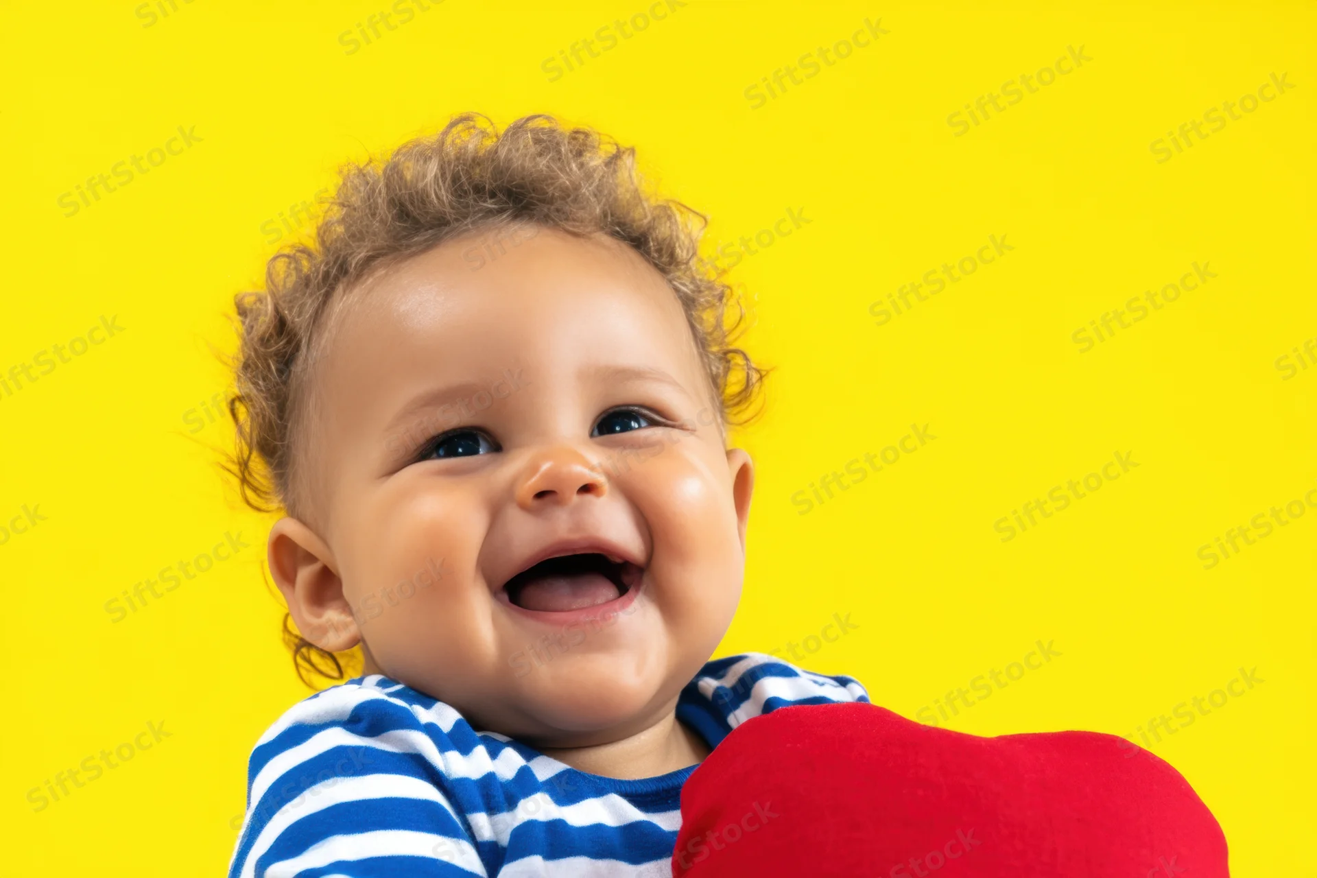 Cheerful baby with curly light brown hair laughing while holding a red heart-shaped plush toy against a yellow background