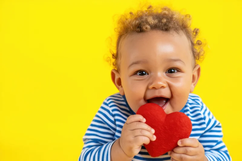 Cheerful baby with curly light brown hair wearing a blue striped shirt holding a red heart toy against a yellow background