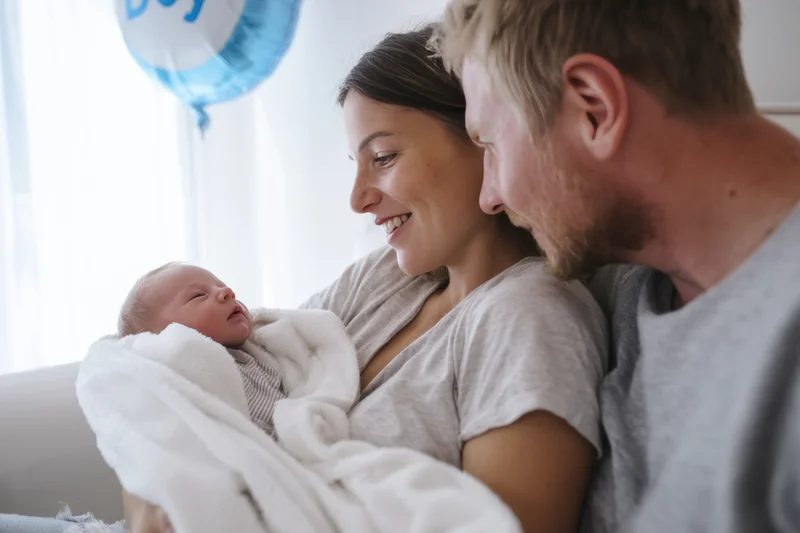 Couple cradling newborn baby wrapped in white blanket on light sofa in bright room