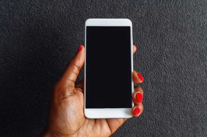 Woman's hand with red nail polish gripping a white smartphone with blank screen against dark textured background