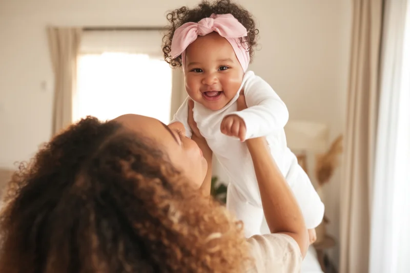 Mother holding and lifting a smiling baby wearing a white onesie and pink headband indoors