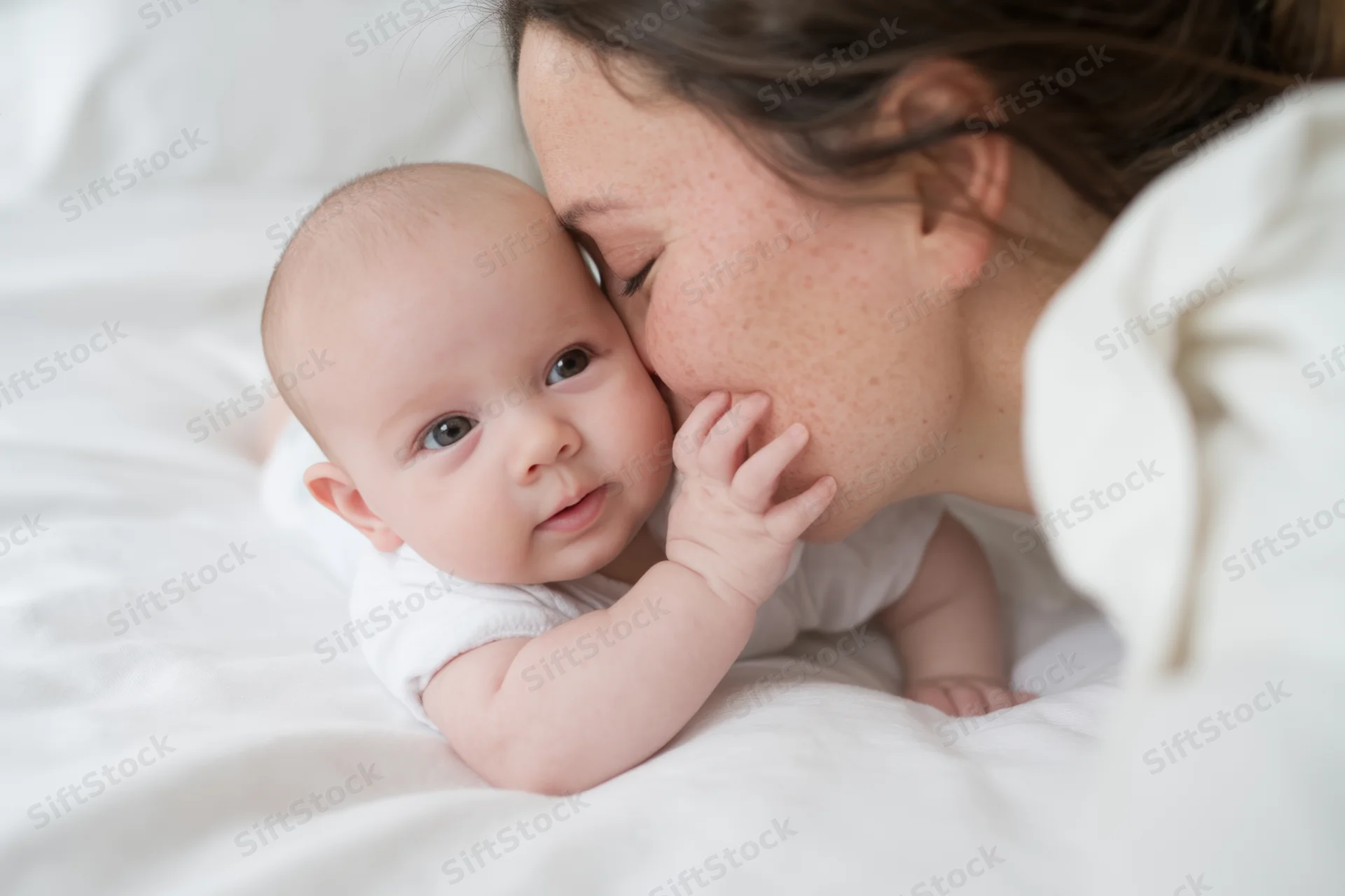 Mother kissing baby on white bed, baby looking at camera, hand touching face