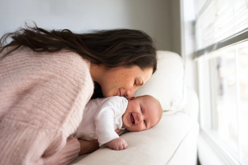 Mother leaning over laughing baby on white couch near window