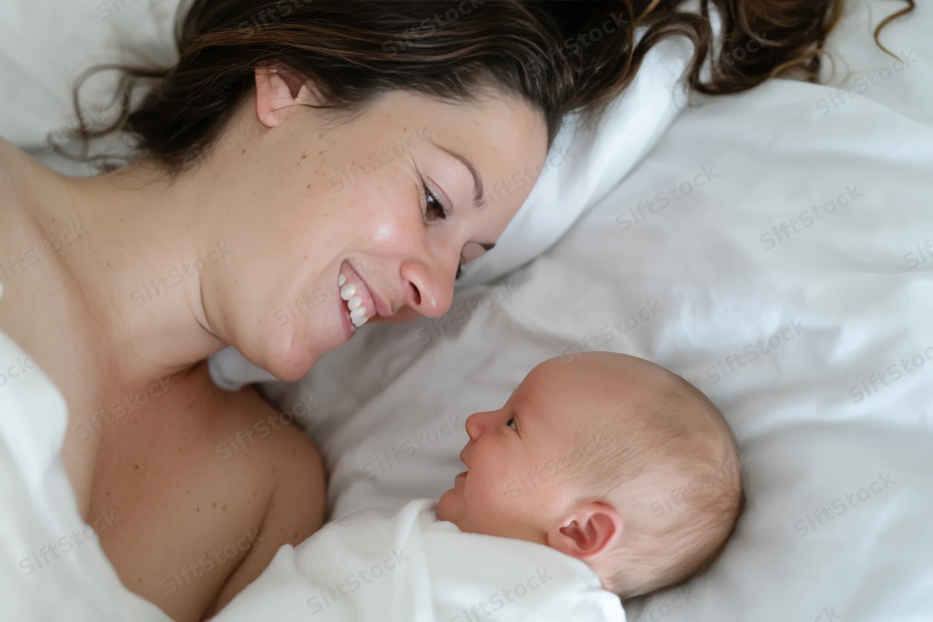 Mother smiling at newborn baby lying beside her on white bed