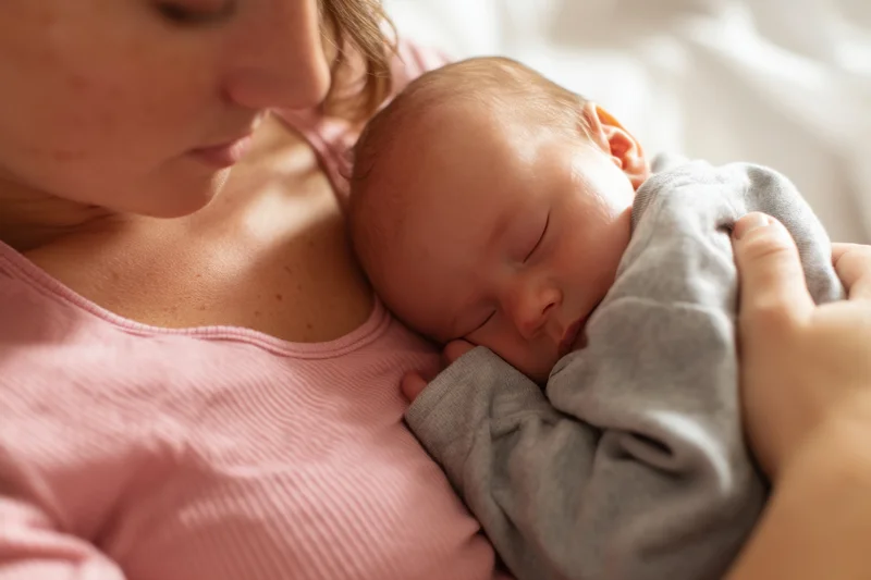 Mother cradling sleeping newborn against her chest in soft indoor light