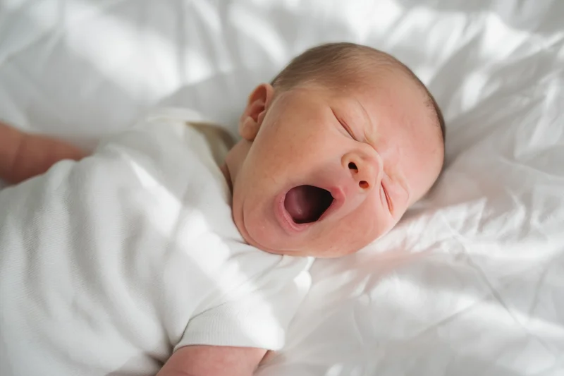 Newborn baby yawning while lying on a white bed with soft bedding