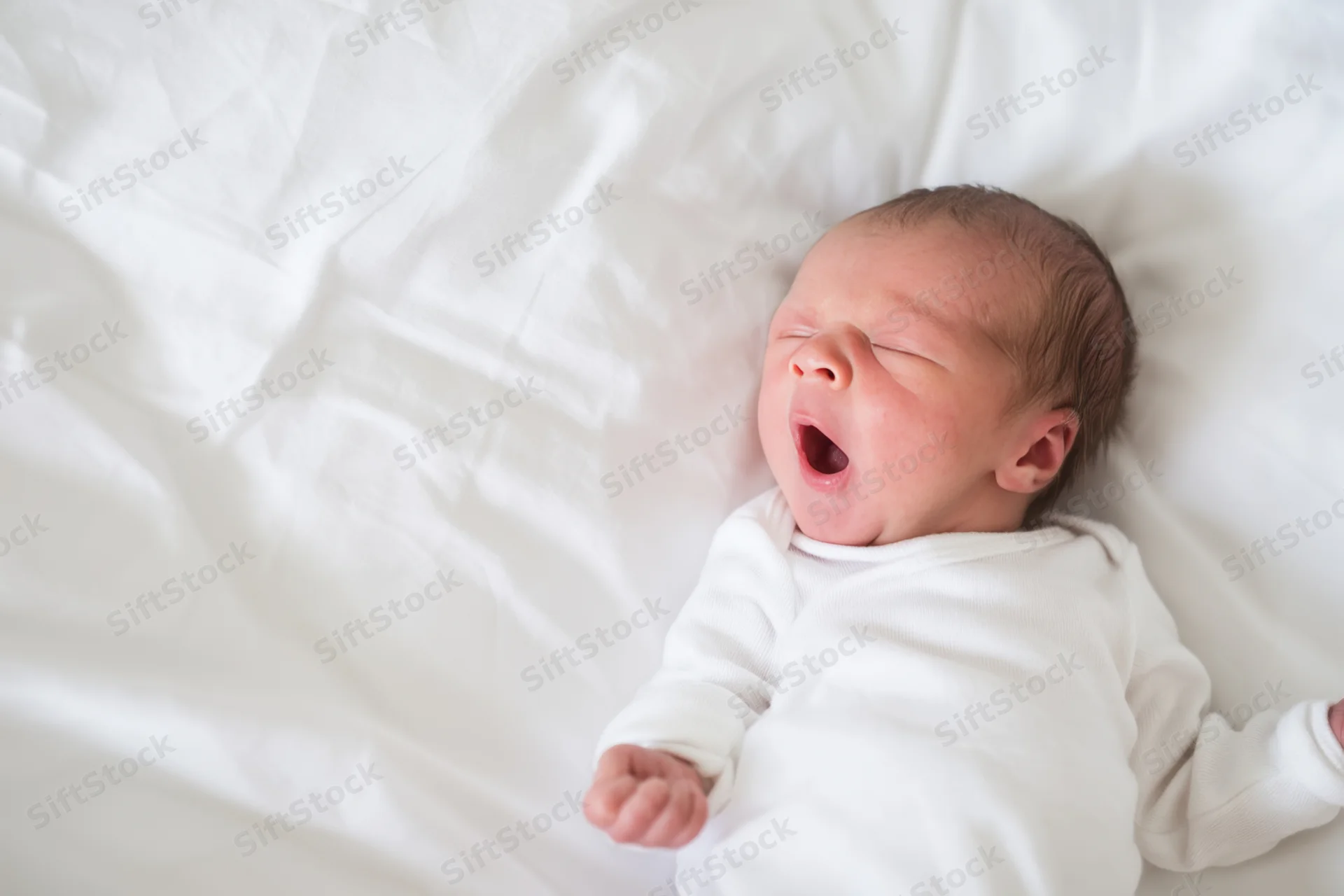 Newborn baby yawning while lying on a white bed blanket