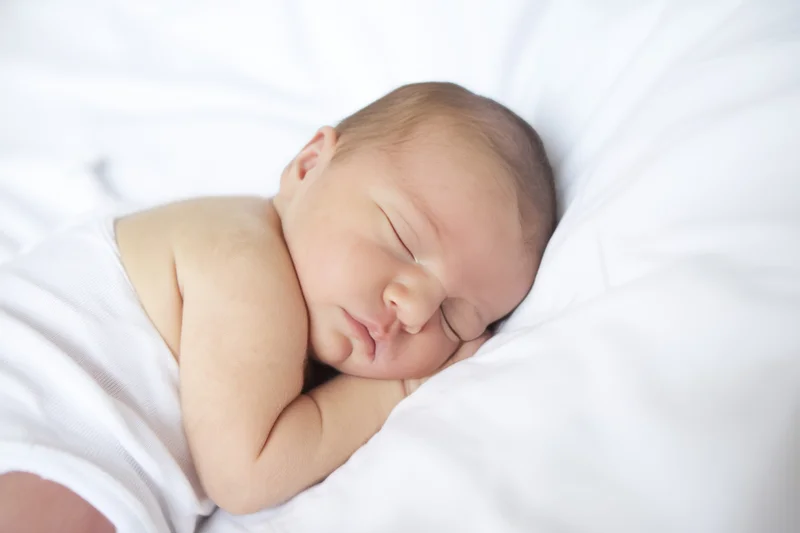 Newborn baby sleeping peacefully on a white blanket with tiny arms folded near face