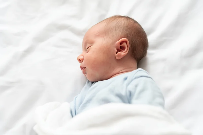 Newborn baby sleeping on white sheets wearing pale blue onesie