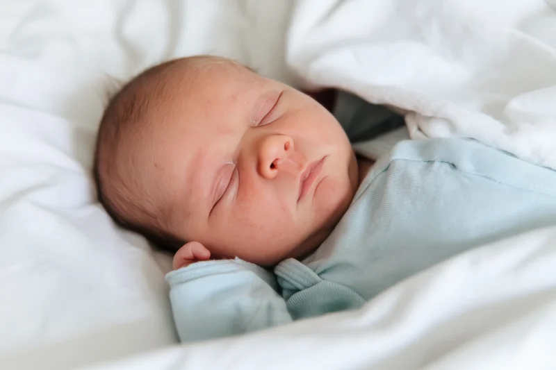 Newborn baby sleeping peacefully on white sheets wearing a pale blue onesie