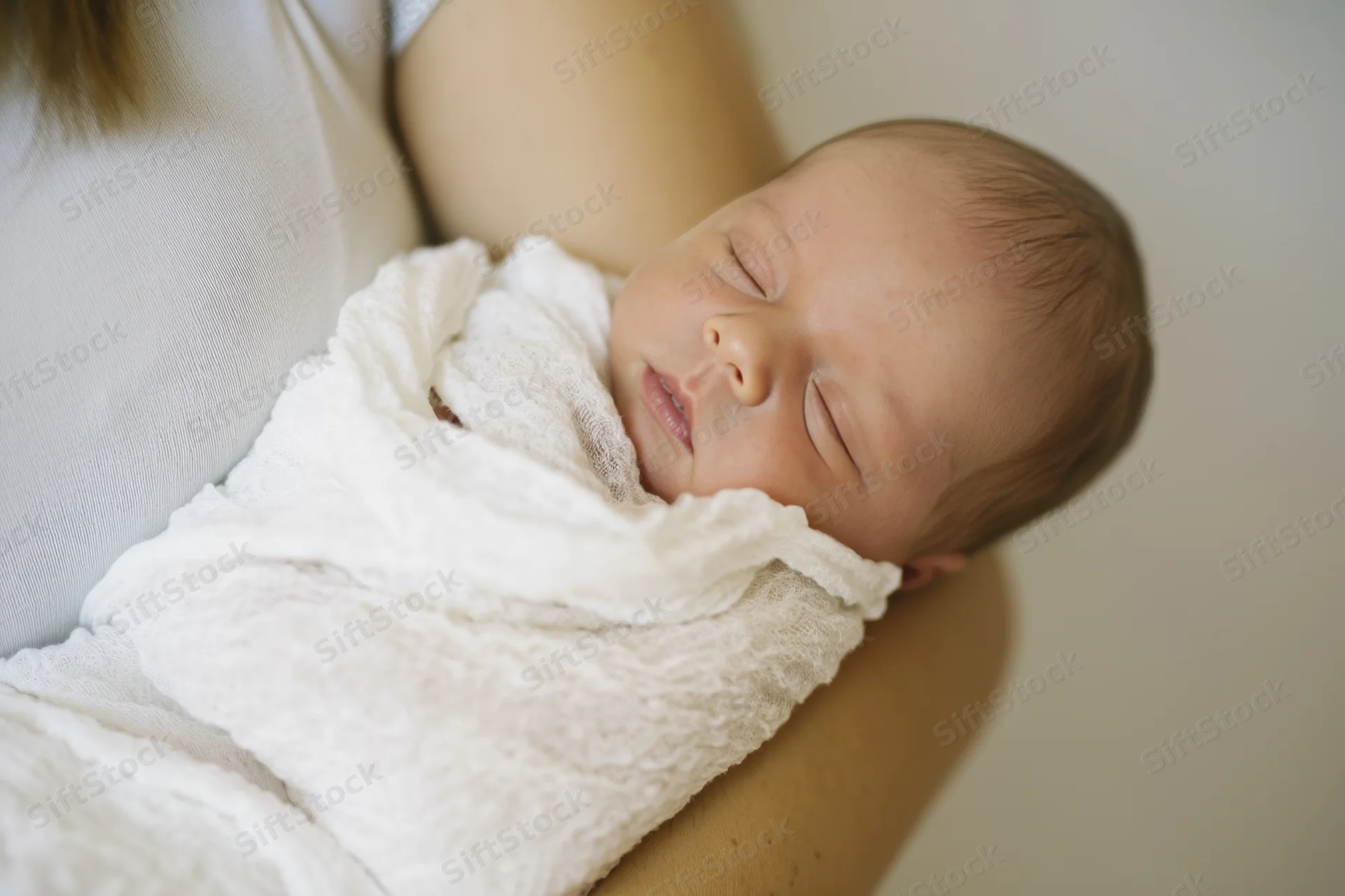Newborn baby sleeping swaddled in white blanket cradled in woman's arms