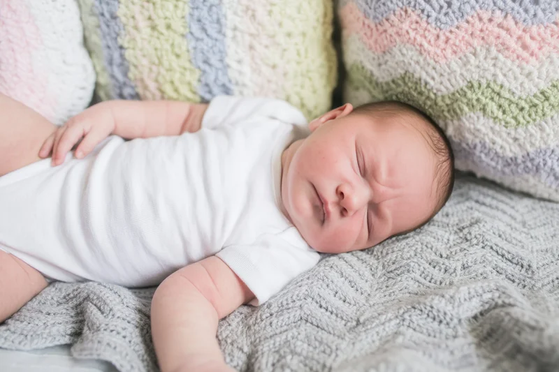 Newborn baby sleeping peacefully on a soft gray knitted blanket with pastel striped cushions in the background