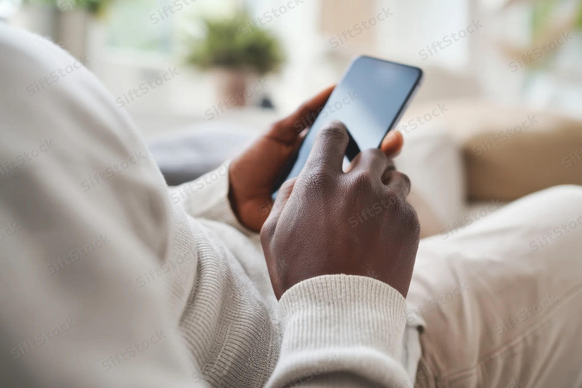 Close-up of a man’s hands using a smartphone while seated on a couch in a living room