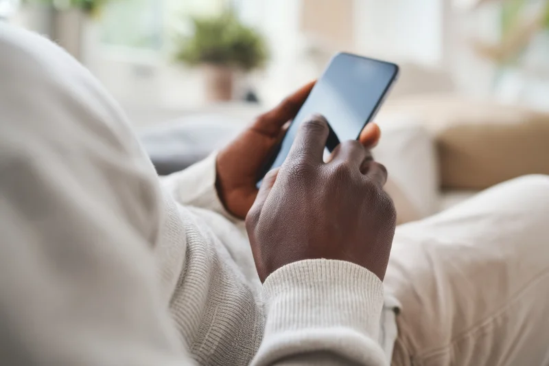 Close-up of a man’s hands using a smartphone while seated on a couch in a living room