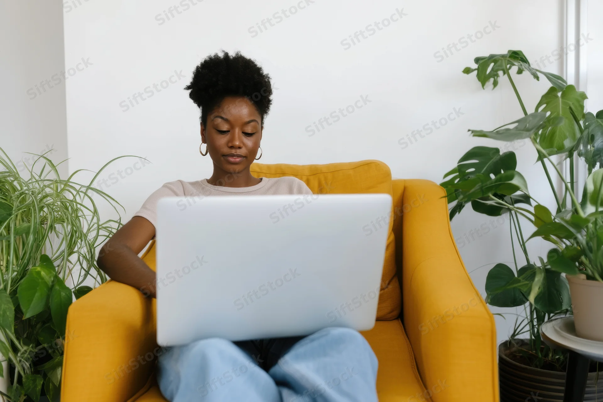 Woman typing on laptop while sitting in yellow armchair surrounded by houseplants